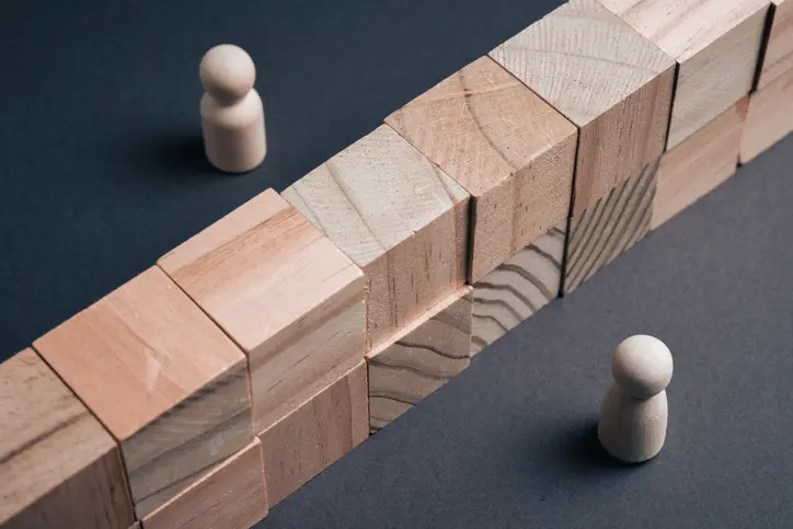 Two wooden peg figures are positioned on opposite sides of a tall wall made of wooden blocks on a dark surface, symbolizing a communication barrier, social divide, or conflict in a business environment