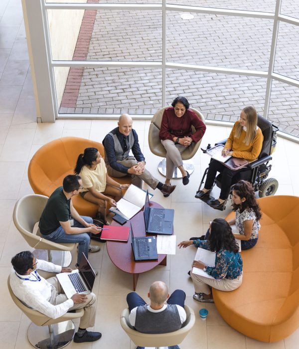 A birds-eye view of a diverse group business meeting. The room is in natural light and the people are of varying ethnicities and ages.